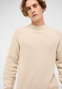 Beiger Strickpullover mit strukturiertem Muster, geripptem Rundhalsausschnitt und langen Raglanärmeln. Der Stoff wirkt weich und leicht.