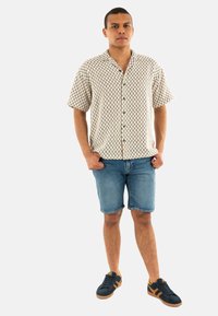Camicia a maniche corte button-up beige con motivo a quadri, abbinata a pantaloni corti di jeans blu. Il modello indossa sneaker scure con dettagli arancioni.