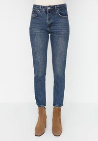 Jeans de mezclilla azul de cintura alta con un ajuste slim, textura desgastada, costuras de cobre y cinco bolsillos, combinados con botas de tobillo de ante marrón.