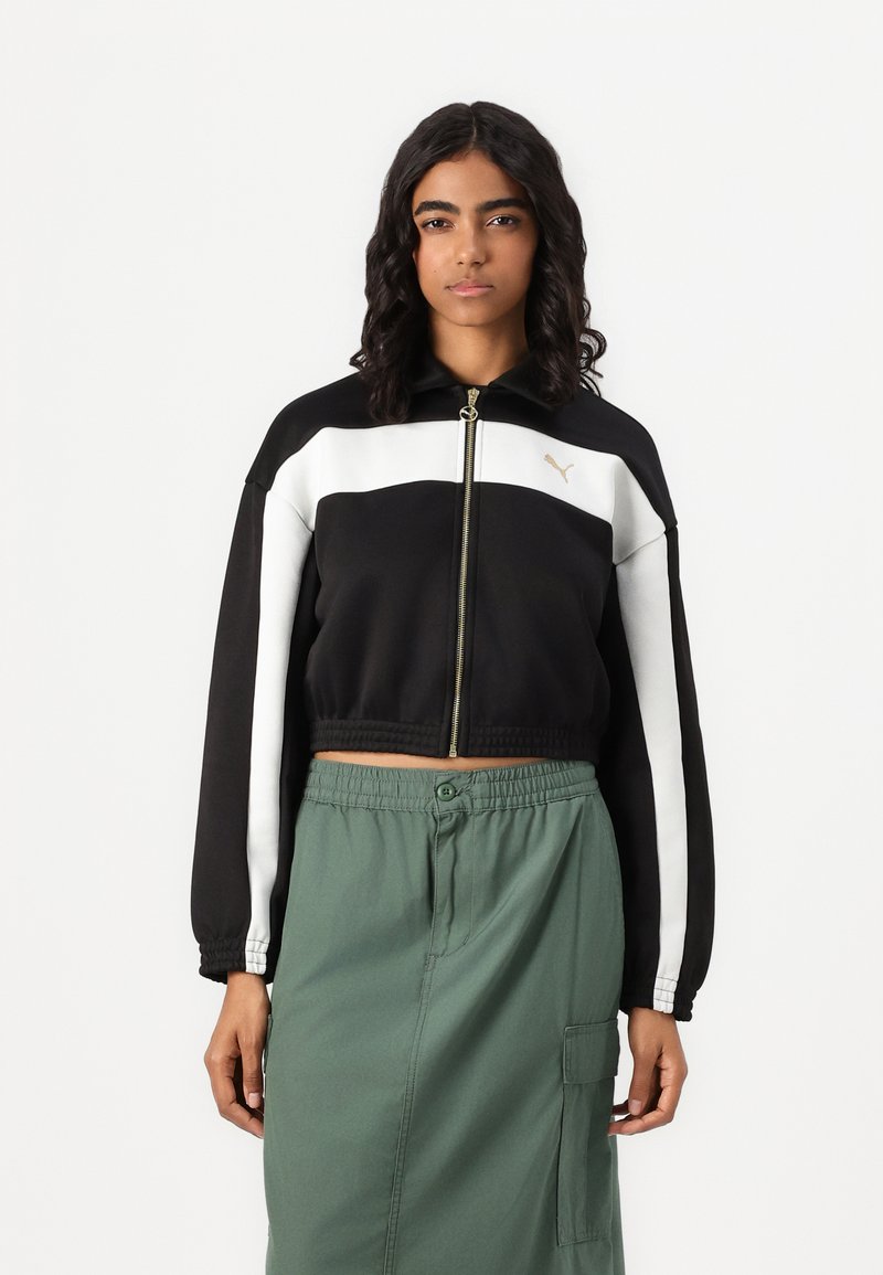 Skraćená čierno-biela bunda s zlatým logom, zapínanie na zips a jemná textura, v kombinácii so zelenými cargo nohavicami s elastickým pásom.
