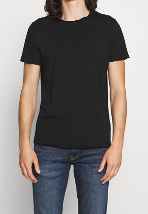 Person iført en helt sort t-shirt med korte ærmer og blå jeans, stående mod en ensfarvet lys baggrund.