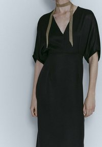 Sort, flydende kjole med V-hals og vide ærmer. Har en tekstureret guldkæde detalje omkring halsen. Glat, mat stof.