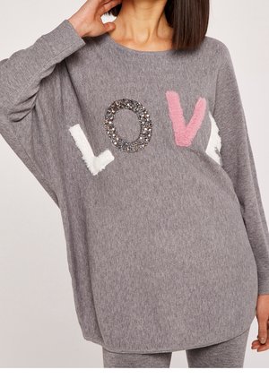 Femme portant un pull gris ample avec les lettres L, O, V, E en textures et couleurs mélangées sur la poitrine.