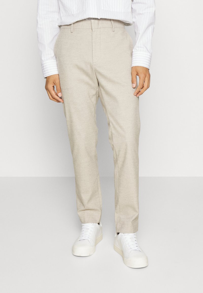 Beige Hosen aus leichtem Stoff mit geradem Schnitt. Kombiniert mit weißen Sneakers und einem kurz geschnittenen, gestreiften weißen Hemd.