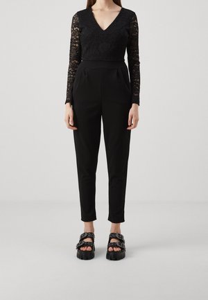 Top de encaje negro de manga larga con escote en V y pantalones negros de cintura alta. Combinado con sandalias negras gruesas con hebillas.