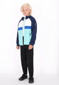 Farvet jakke i aqua, hvid og marineblå, med lynlås foran og hætte. Kombineret med sorte bukser og mørke sneakers. Glat tekstur.