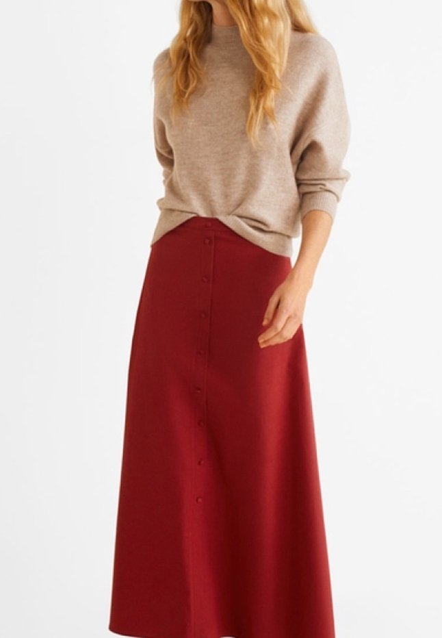 Femme portant un pull beige à manches longues et une jupe midi rouge taille haute boutonnée devant, debout devant un fond blanc.