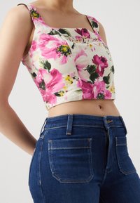 Blommig crop top med rosa och gröna mönster på en krämfärgad bakgrund, med fyrkantig halsringning och breda band, parat med mörkblå jeans.