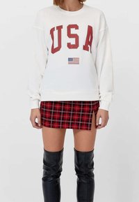 Sudadera blanca con "USA" en letras rojas y una pequeña bandera americana. Combinada con una falda corta de cuadros rojos y botas negras hasta la rodilla.