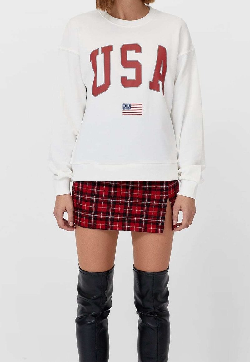 Sudadera blanca con "USA" en letras rojas y una pequeña bandera americana. Combinada con una falda corta de cuadros rojos y botas negras hasta la rodilla.