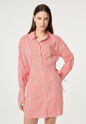 Vrouw draagt een rood-wit verticale gestreepte jurk met knopen, lange mouwen en grote oorringen, staand tegen een witte achtergrond.