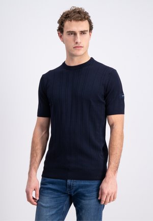 Man with curly hair wearing a navy short-sleeve ribbed knit shirt and blue jeans, standing against a plain light background.