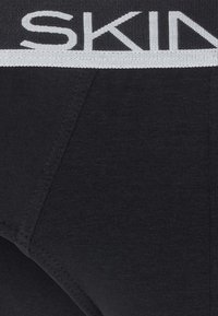 Slip di cotone neri con un elastico intrecciato che presenta la parola "SKIN" in bianco. Il tessuto è morbido e a coste, con dettagli di cucitura puliti.