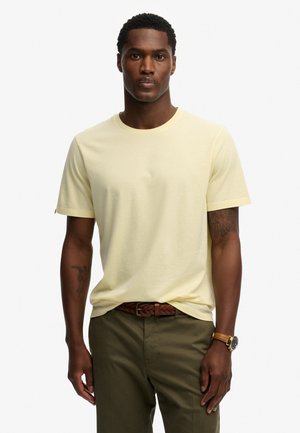 Man draagt een lichtgeel T-shirt met korte mouwen, olijfgroene broek met een gevlochten bruine riem en een polshorloge met een bruine leren band.