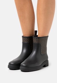 Jambes portant des bottes de pluie noires à mi-mollet avec des dessus en tissu à motifs marron arborant un logo "CK" répété sur fond blanc.