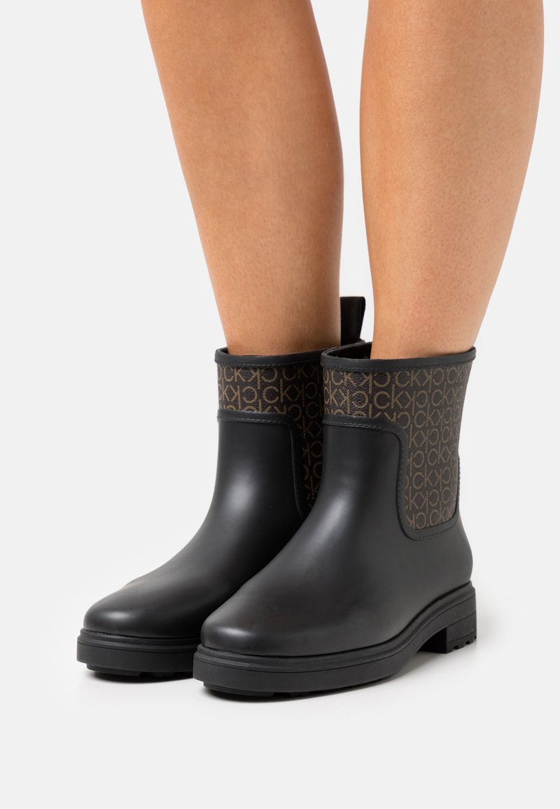 Jambes portant des bottes de pluie noires à mi-mollet avec des dessus en tissu à motifs marron arborant un logo "CK" répété sur fond blanc.