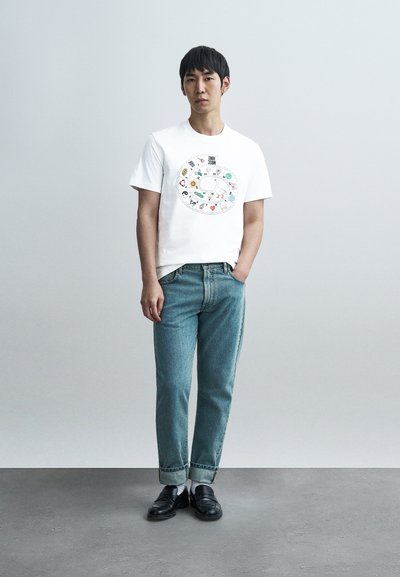 Λευκό βαμβακερό t-shirt με πολύχρωμο γραφικό σχέδιο. Συνδυασμένο με τζιν παντελόνι ανοιχτού μπλε και μαύρα slip-on παπούτσια.