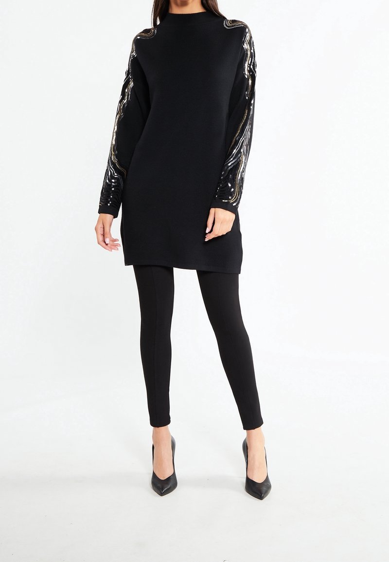 Donna che indossa una tunica nera a maniche lunghe con strisce di paillettes argento e oro sulle maniche, leggings neri e décolleté neri a punta.
