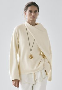 Beige knit sweater with an asymmetrical design, layered over a white shirt. Gold pendant on a black cord adds a unique detail.