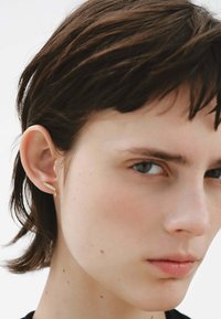 Pendiente de oro con un diseño delgado y curvado, adornado con una pequeña piedra decorativa. El modelo tiene el cabello corto castaño y piel clara. Estilo simple y minimalista.