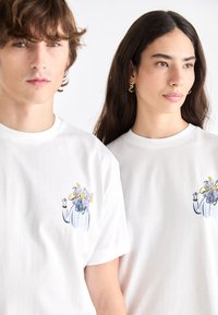 Hvide bomulds t-shirts med et blåt og gult blomsterdesign på venstre bryst, rund hals og korte ærmer.
