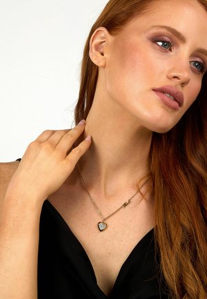 Collana in oro con un pendente a forma di cuore con una pietra scintillante al centro, indossata su una maglietta nera.