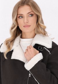 Schwarze Kunstlederjacke mit weißem Schafslederkragen und Manschetten. Verfügt über einen Reißverschluss und eine Metall-Schnallenakzentuierung. Weiche Textur und warmes Design.