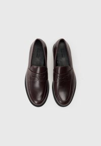 Κάστανες δερμάτινες μοκασίνες με λείος φινίρισμα, διακοσμημένες με ραμμένο διακοσμητικό στοιχείο κατά μήκος του πάτου και μαύρη ελαστική σόλα.