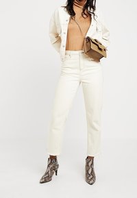 Žena oblečená v béžovém roláku, off-white bundě a džínech, hadích kotníkových botách a hnědé kabelce přes rameno s řetízkovým popruhem.