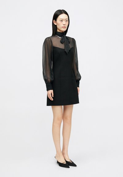 Μαύρο μίνι φόρεμα με διαφάνες φουσκωτές μανσέτες, λεπτομέρεια με κορδόνι στο λαιμό και εφαρμοστό σχέδιο. Λείο ύφασμα, συνδυασμένο με μαύρες μυτερές γόβες.