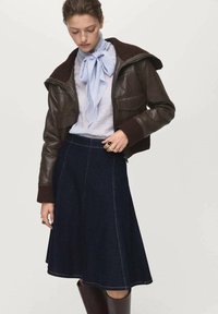 Veste en cuir marron avec une capuche, écharpe bleu clair, chemise blanche et jupe ligne A en denim foncé. Bottes marron jusqu'aux genoux au design épuré.