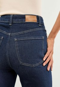 Tumedad denim teksad on sobivustega, pruuni nahk riiulpesaga ja kaks tagumist taskut kontrastse õmblusega.