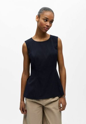 Kvinne med kort hår iført en marineblå ermeløs topp med pinstripes og beige bukser mot en ensfarget hvit bakgrunn.