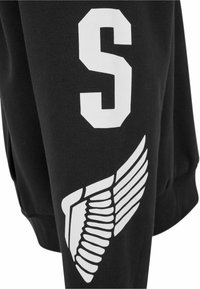 Sudadera negra con una gran "S" blanca en la manga y un gráfico de ala blanca, ambos diseñados en estilos distintos y audaces.