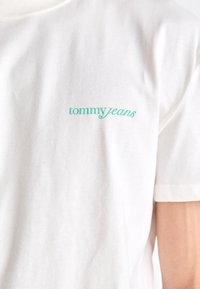 White cotton t-shirt featuring a turquoise embroidered logo reading "tommy jeans" on the left chest area. Classic crew neck design.