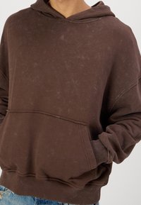 Sudadera marrón hecha de un tejido suave y texturizado. Cuenta con un bolsillo frontal, un corte holgado y una capucha con cordón. Los detalles desgastados añaden un interés visual.