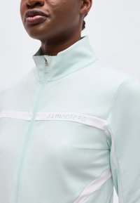 Κοντινό πλάνο ενός ατόμου που φοράει ανοιχτό μπλε ζακέτα με φερμουάρ, λευκές ρίγες και το λογότυπο "J.LINDEBERG" στο στήθος.