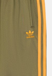 Olivno zeleni atletski hlače z oranžnimi stranskimi črtami. Imajo adidas logo v oranžni vezenini. Gladka tekstura z elastiko v pasu.