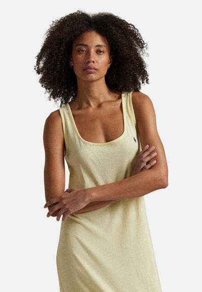 Femme aux cheveux bouclés portant une robe sans manches jaune pâle, debout les bras croisés devant un fond clair uni.