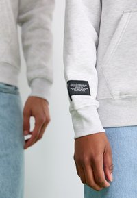 Sweatshirt gris clair avec un poignet côtelé. Il présente une étiquette en tissu noir sur la manche avec un texte imprimé. Une jupe en denim bleu est visible à l'arrière-plan.