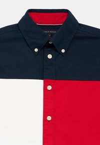 Πουκάμισο με κοντά μανίκια και γιακά, με μπλοκ χρωμάτων σε ναυτικό μπλε, κόκκινο και λευκό, κλείσιμο με κουμπιά και αντίθετο κόκκινο εσωτερικό γιακά.