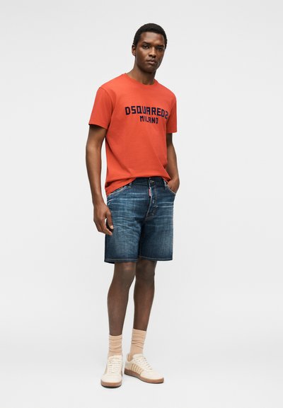 Άνδρας που φοράει κόκκινο μπλουζάκι «DSQUARED2 MILANO», μπλε τζιν σορτς, μπεζ κάλτσες και λευκά αθλητικά παπούτσια, στέκεται με το ένα χέρι στην τσέπη.