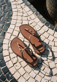Tongs marron avec des clous dorés sur un pavement en pierre mosaïque noir et blanc, projetant des ombres sous la lumière naturelle du soleil.