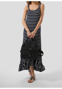 Sac à dos noir avec une poignée supérieure et un rabat, comportant deux poches avant et une quincaillerie en argent. Le modèle porte une robe maxi rayée et des sandales.