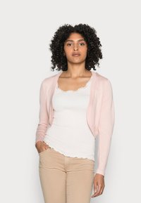 Light pink cardigan with long sleeves, open front design. Worn over a white tank top with a scalloped neckline. Smooth knit texture.