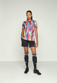 Maglietta da calcio a maniche corte e multicolore con design geometrico, abbinata a pantaloncini neri e calzettoni neri al ginocchio con strisce bianche.