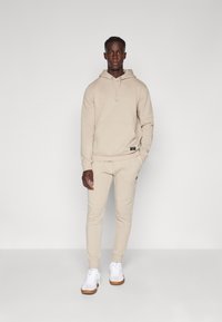 Sweatshirt bege com capuz e bolso frontal, calças de treino ajustadas a combinar, e tênis brancos com solas castanhas. Textura de tecido suave.