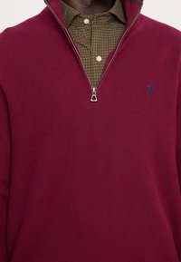 Burgunderfarbener Viertelzip-Pullover mit strukturiertem Strickmuster und glattem Reißverschluss, ausgestattet mit einem gestickten blauen Logo auf der Brust.