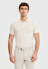 Beżowa koszulka polo wykonana z gładkiego materiału, z klasycznym kołnierzykiem i krótkimi rękawami, zestawiona z jasnobeżowymi spodniami. Prosty design.