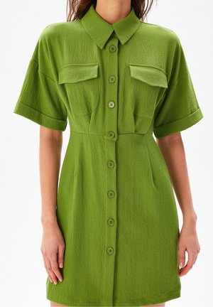 Frau trägt ein strukturiertes, olivgrünes, kurzärmeliges Hemdkleid mit durchgehender Knopfleiste, zwei Brustklappen und Kragen.